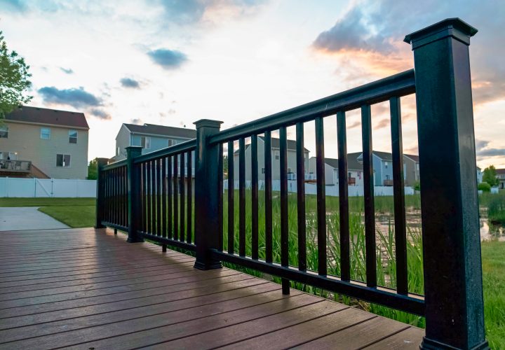 Bridge over a pond with multi storey homes and cloudy sky in the background. The structure has a brown wood deck and black guardrail.